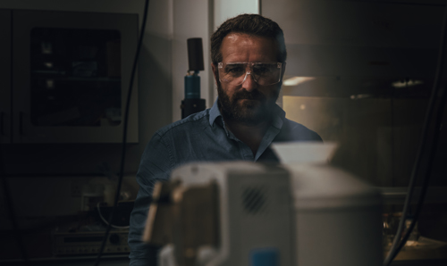 Man in goggles and blue shirt stood in dimly lit room